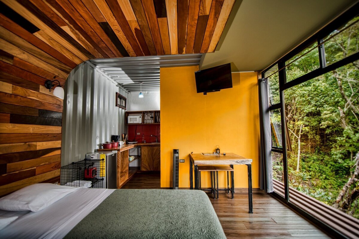 A compact room with a bed, a small kitchenette, a dining area with a table and chairs, and a large window offering a view of greenery.