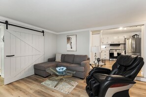 Una sala moderna con un sofá seccional gris, una mesa de centro de vidrio, un sillón de masaje y una cocina al fondo.