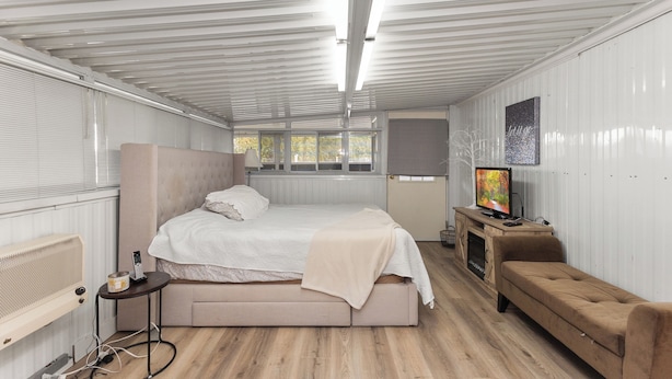 A compact, well-lit room with a bed, a bench, a TV, and a small table.
