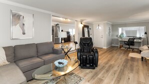 A modern living room with a gray sofa, a glass coffee table, a massage chair, and a desk with a computer.