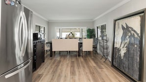 A modern kitchen with a stainless steel refrigerator, wooden flooring, a dining area with a table and chairs, and a large abstract painting on the wall.
