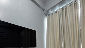 En stue med et fladskærms-tv monteret på en træhylde, en grå sofa med pyldepuder og et vindue med lysegrå gardiner.