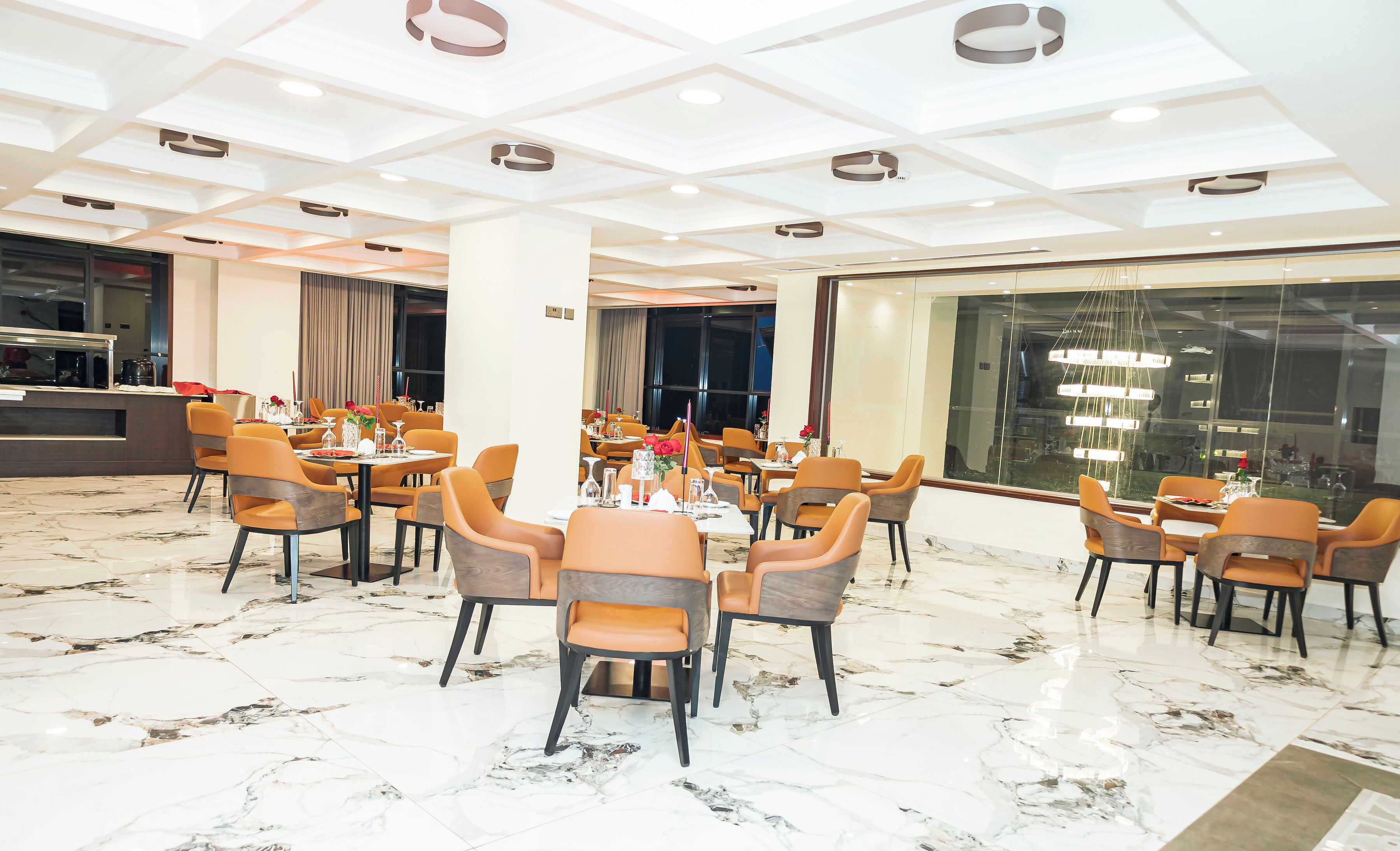 A spacious dining area with marble flooring and modern orange chairs. Tables are set with red candles and floral centerpieces.