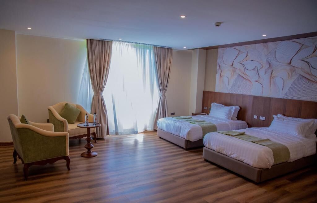 Ein Hotelzimmer mit zwei Betten, einem grünen Sessel, einem kleinen runden Tisch und einem großen abstrakten Wandbild.