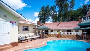 A swimming pool with lounge chairs and umbrellas, a warning sign, and a building with a tiled roof. - THE MAVVEL (Mutunduri)