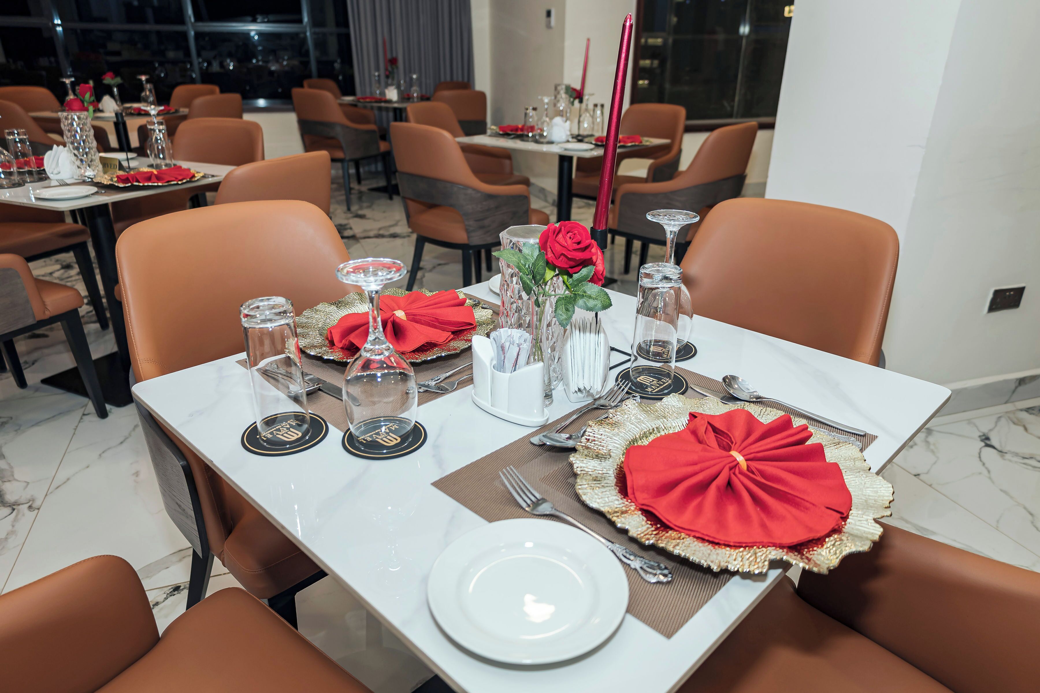 A dining area with tables set for two, featuring white plates, red napkins, and decorative glassware.