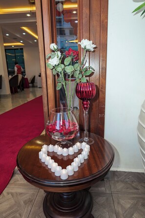 A table with a heart-shaped arrangement of candles, a glass vase with white roses, and a red glass vase with a single rose. - THE MAVVEL (Mutunduri)