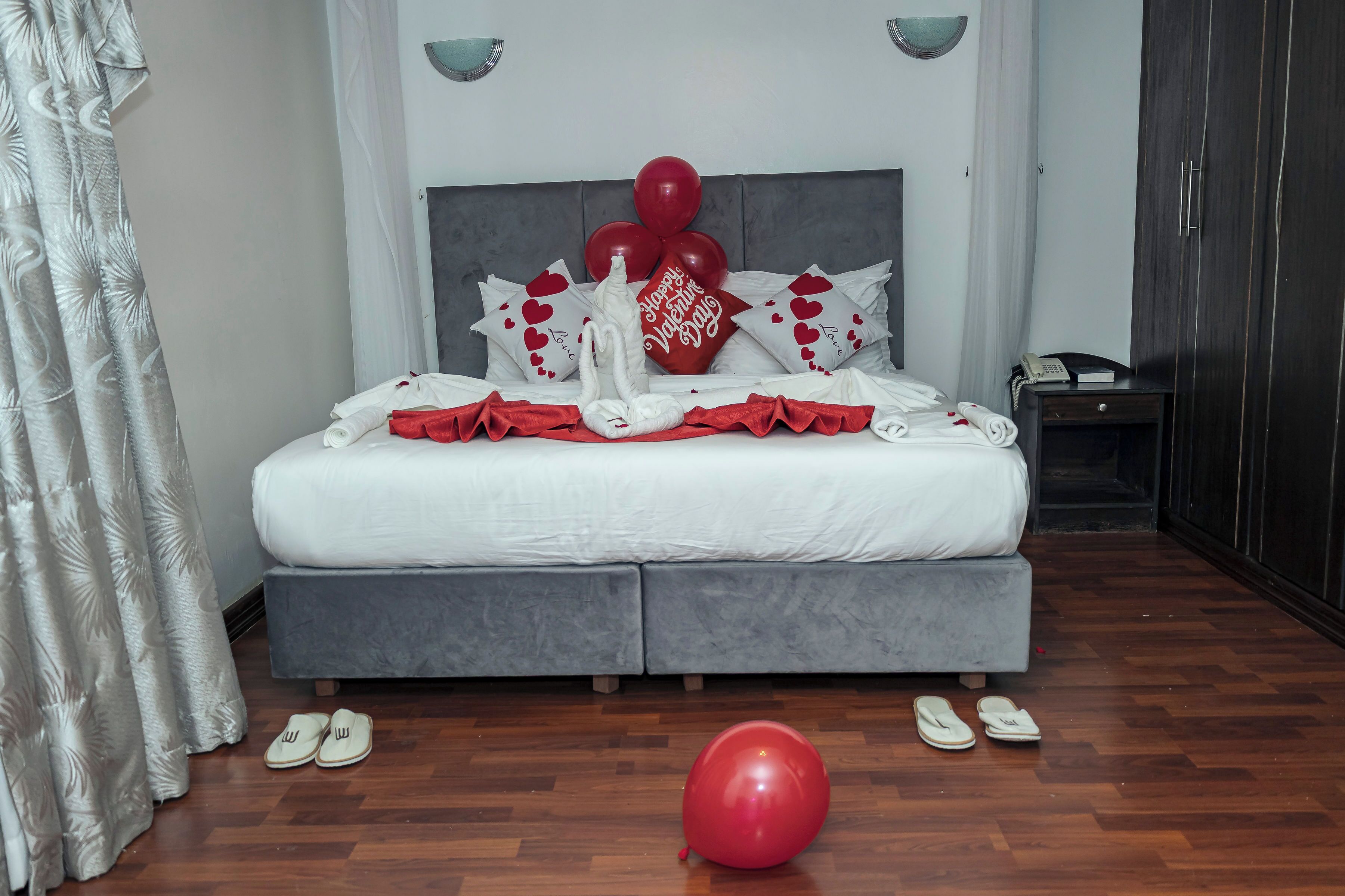Un lit bien fait, avec une literie blanche, une tête de lit grise et des oreillers rouges et blancs. On peut voir des ballons rouges et une décoration en forme de cygne sur le lit.