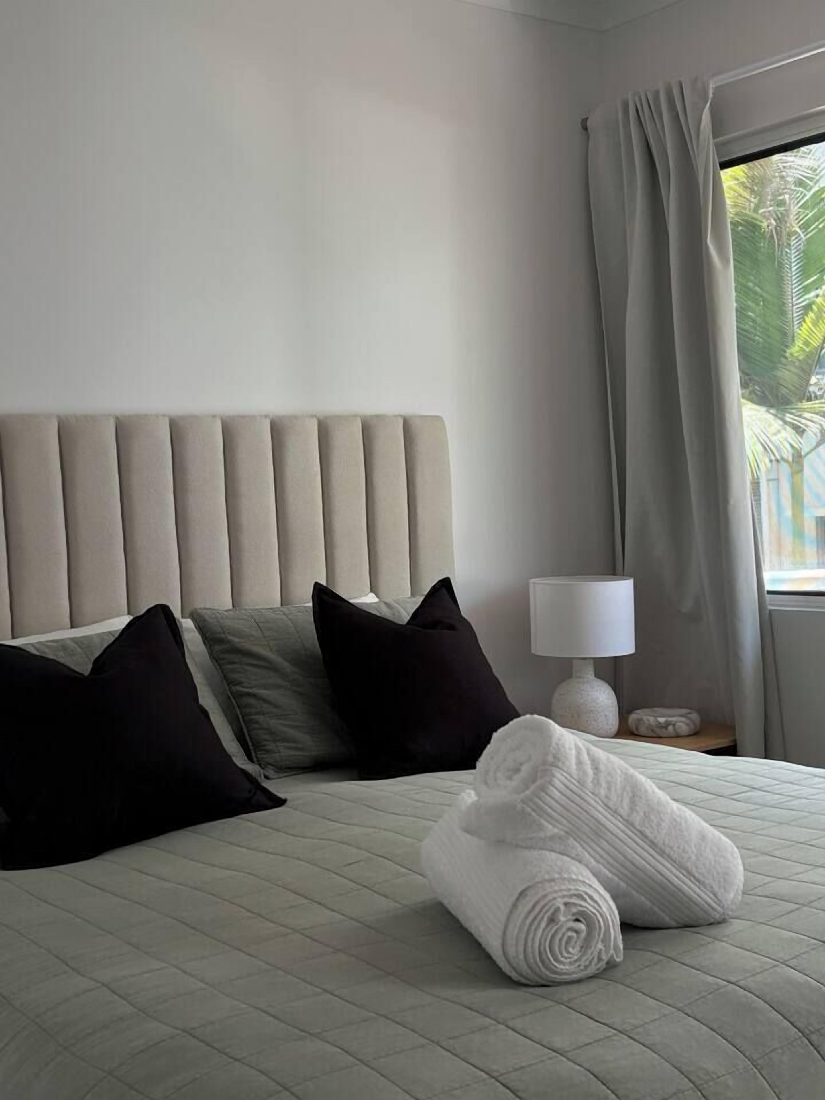 Een hotelkamer met een bed, kussens, een opgerolde handdoek, een lamp en een raam met gordijnen.