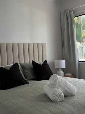 A hotel room with a bed, pillows, a rolled towel, a lamp, and a window with curtains.