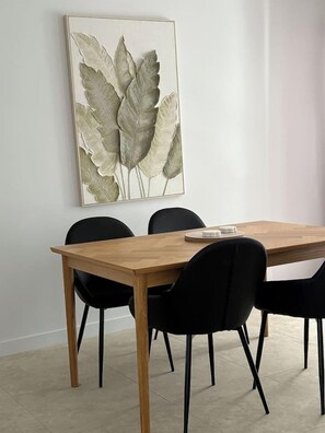 Un comedor con una mesa de madera y cuatro sillas negras. Un cuadro con hojas está colgado en la pared.