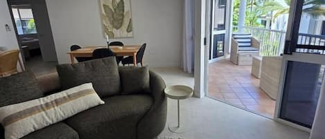 Un salon comprenant un canapé, une table basse et un tapis à motifs. On y trouve également un coin repas avec une table et des chaises, ainsi qu’une vue sur un balcon aménagé avec du mobilier d’extérieur.