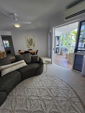 Una sala de estar con sofá, una mesa de centro y un tapete estampado. Hay un comedor con mesa y sillas, y vistas a un balcón con mobiliario exterior.