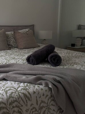 Un lit bien fait, avec une couvre-lit à motifs et des oreillers, une couverture enroulée et une table de chevet sur laquelle se trouvent une lampe et un objet décoratif.