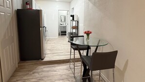 A narrow hallway with a dining area featuring a glass table and chairs, a refrigerator, and a kitchen in the background.