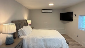 A bedroom with a bed, nightstands, a lamp, an air conditioning unit, a TV, and a window.