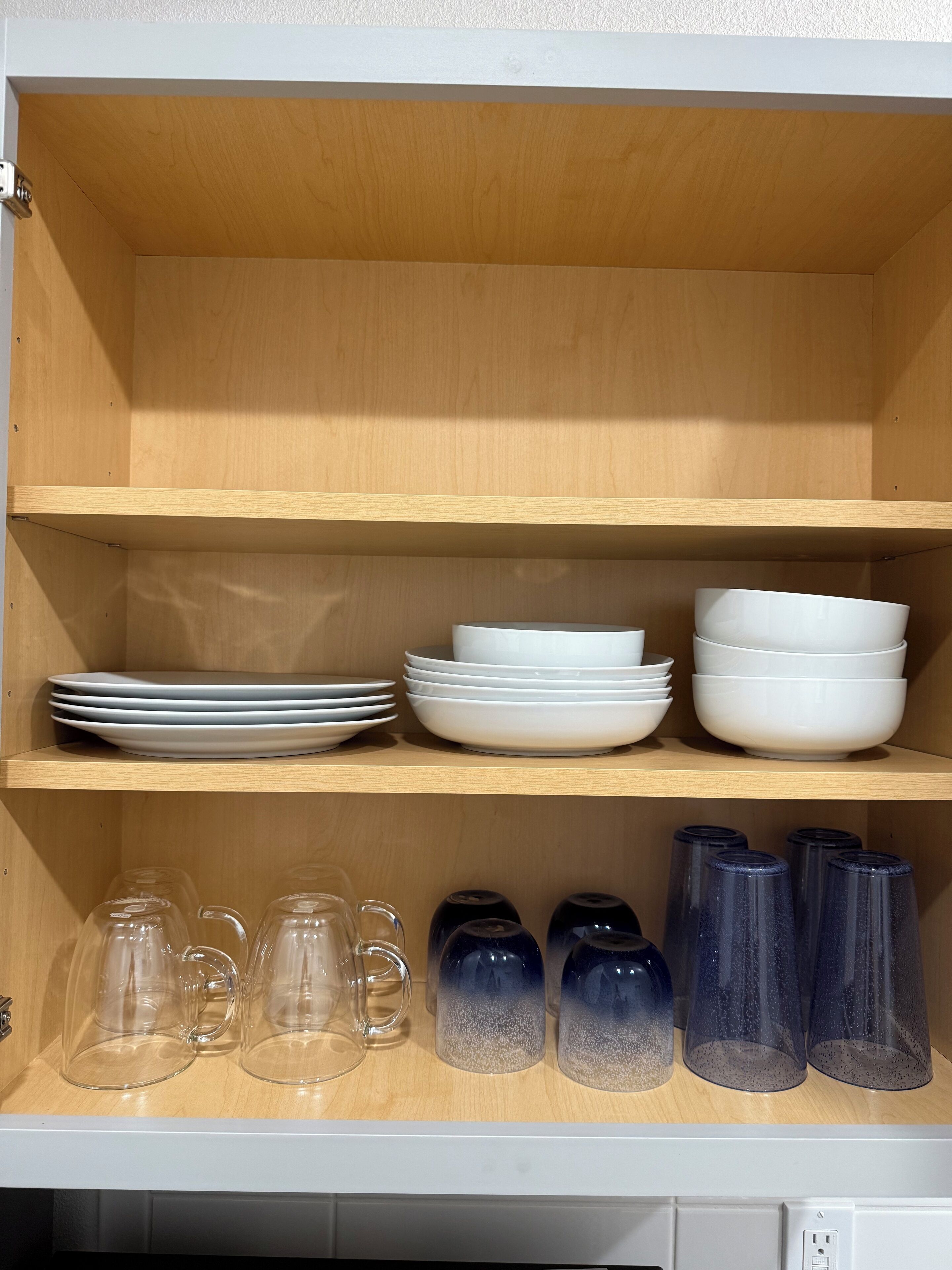 A wooden shelf unit with white plates and bowls stacked on the top shelf and clear glass mugs and tumblers on the bottom shelf.