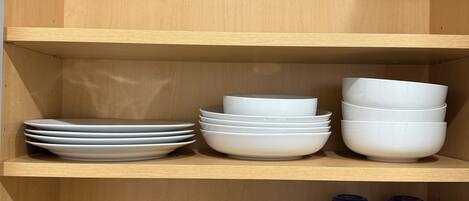 A wooden shelf unit with white plates and bowls stacked on the top shelf and clear glass mugs and tumblers on the bottom shelf.