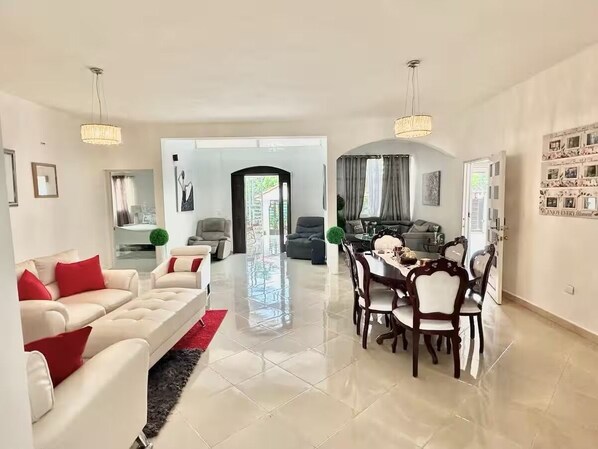 A spacious living room with a dining area, featuring a large window, a chandelier, and a dining table with chairs.