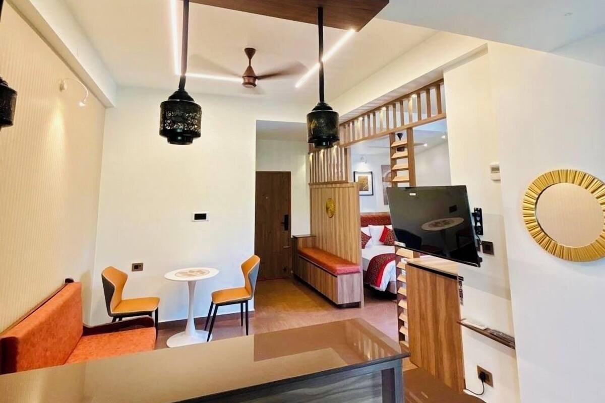 A modern hotel room with a wooden ceiling, a dining area with orange chairs, a TV, and a circular wall decoration.