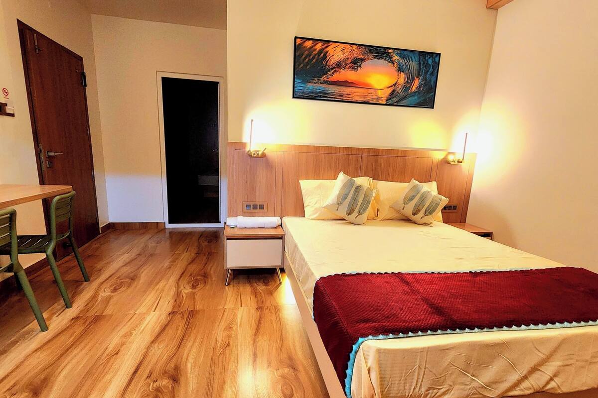 A hotel room with a wooden floor, a bed with a red blanket, a nightstand, a desk with a chair, and a painting of a sunset.