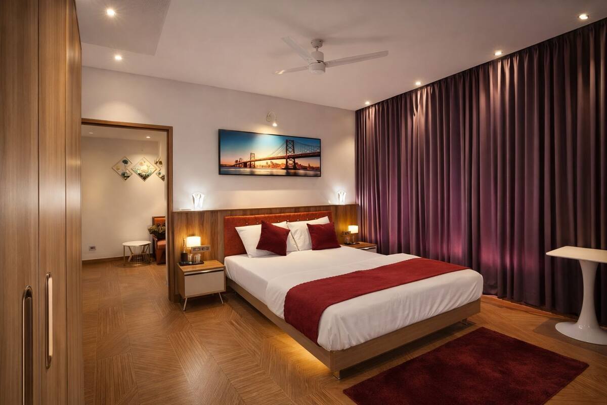 A hotel room with a large bed, wooden flooring, a ceiling fan, a painting of a bridge, and a view of a cityscape.
