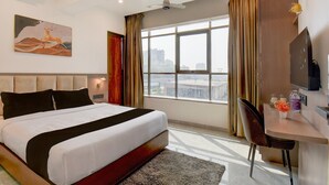 Ein Hotelzimmer mit einem großen Bett, einem Schreibtisch mit Stuhl, einem Flachbildfernseher, einem Fenster mit Vorhängen und einem Bild an der Wand.