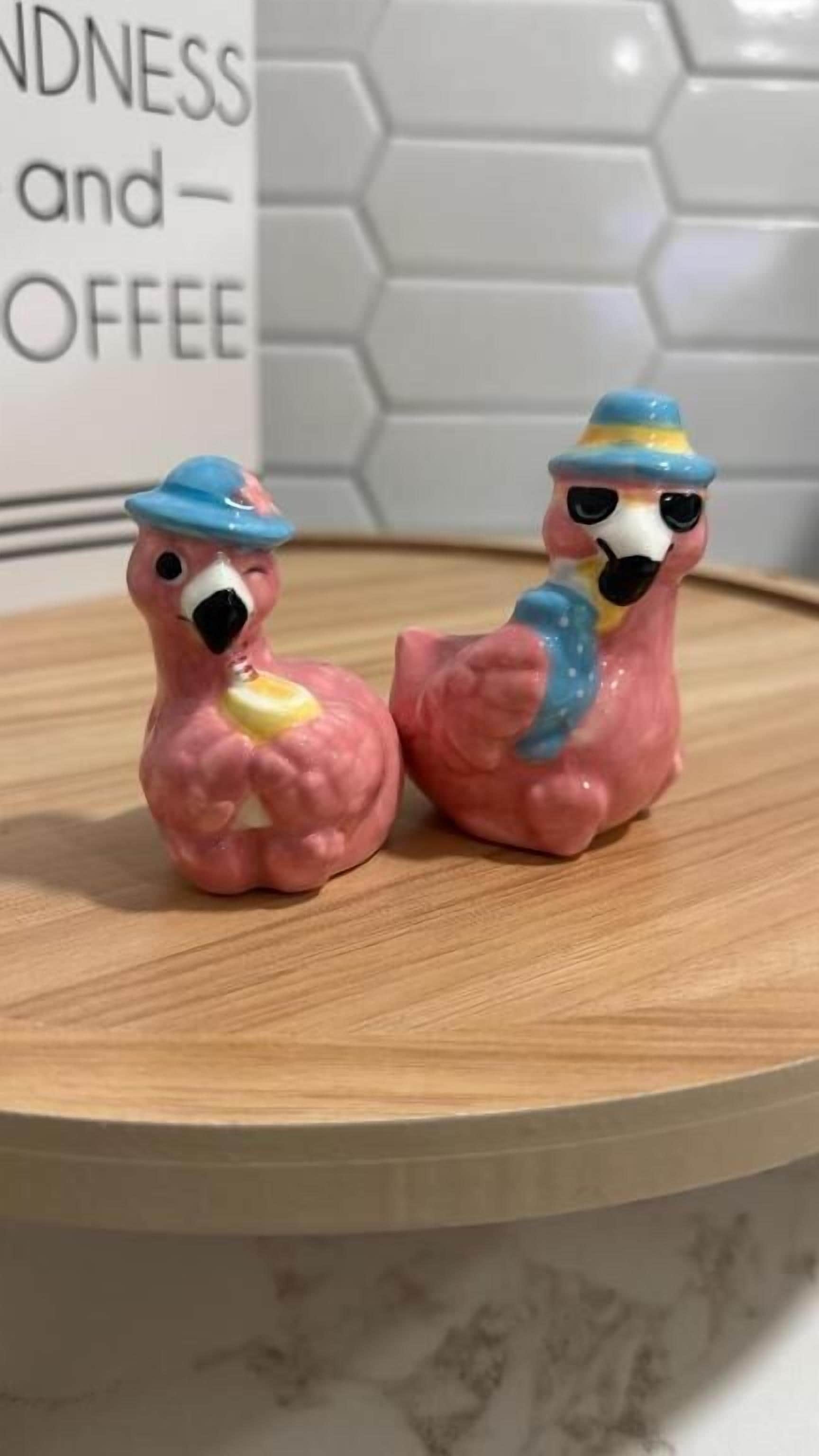 Two pink flamingo figurines on a wooden surface with a hexagonal tile wall in the background.