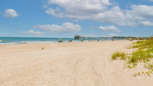Uma praia com um cais que se estende para o oceano, um pequeno edifício perto da água e céu claro.