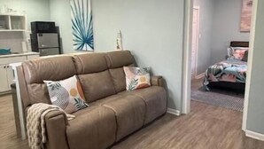 Sala de estar com sofá de couro marrom, área de cozinha com armários e eletrodomésticos, e quarto com cama e janela.
