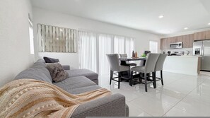 Ein modernes Wohnzimmer mit einem grauen Sofa, einem Esstisch mit Stühlen und einer Küchenzeile inklusive Mikrowelle und Kühlschrank.