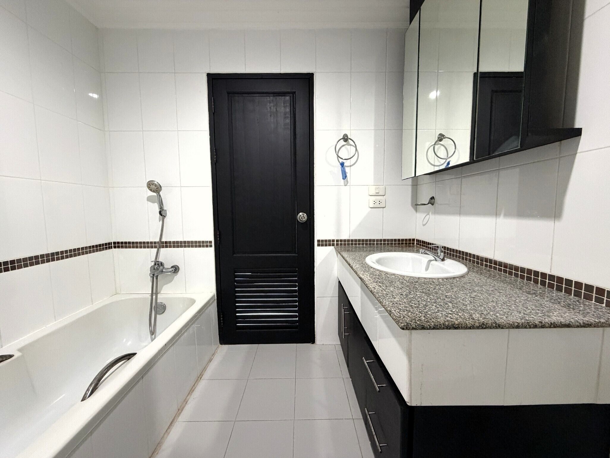 A modern bathroom with a black door, white tiles, a bathtub, a sink, and a mirror.