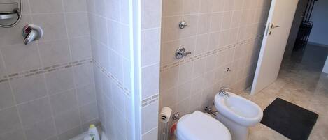 A bathroom with a glass shower enclosure, two toilets, a bidet, and a blue mat on the floor.