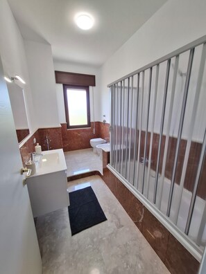 Une salle de bain avec un lavabo blanc, des toilettes, une douche avec une porte en verre et une fenêtre avec un cadre marron.