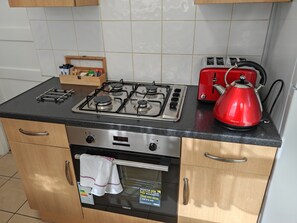 A kitchen with a gas stove, a red kettle, a toaster, and a wooden cabinet with a towel hanging over the oven door. - Apartment With Private entrance, parking & Garden, few minutes to Town Centre (Borehamwood)