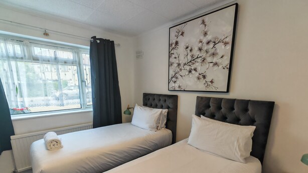 Habitación de hotel con dos camas, un radiador, una ventana con cortinas, un cuadro colgado en la pared y una mesita de noche con lámpara.