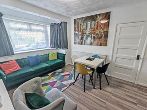 En moderne stue med en grøn sofa, en hvid lænestol, et spisebord med stole og en farverig måtte.