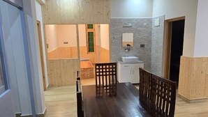 A dining area with a wooden table and chairs, a kitchen area with a sink and cabinets, and a wooden floor. - Aashvika Homes (Manali)