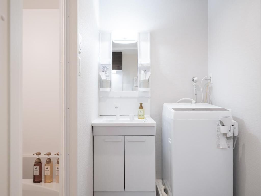 A small, white bathroom with a mirror, sink, and toilet. There are toiletries on the sink and a towel dispenser on the wall.