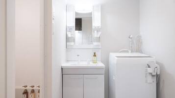 A small, white bathroom with a mirror, sink, and toilet. There are toiletries on the sink and a towel dispenser on the wall.