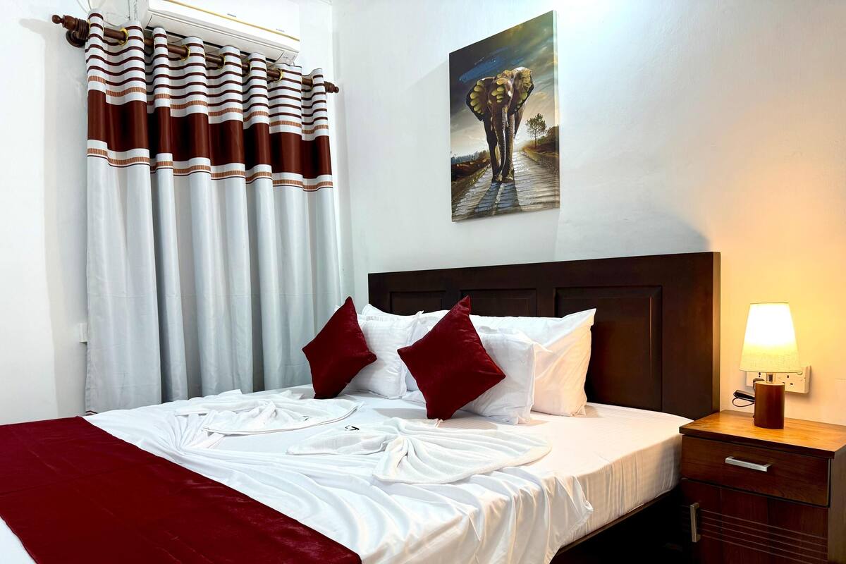 A neatly made bed with white linens and red pillows, a wooden nightstand with a lamp, and striped curtains.