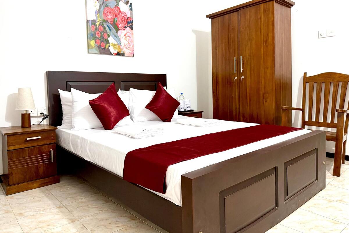 A neatly made bed with white and red bedding, a wooden nightstand with a lamp, a wooden wardrobe, and a wooden chair.
