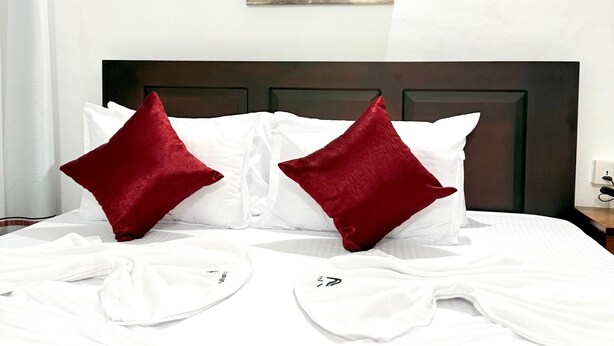 A neatly made bed with white linens and red pillows, a painting of elephants on the wall, and a window with curtains. - Nishi Villa (Negombo)