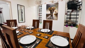A dining area with a wooden table set for four, surrounded by chairs, with artwork on the walls and a window with blinds. - Nishi Villa (Negombo)
