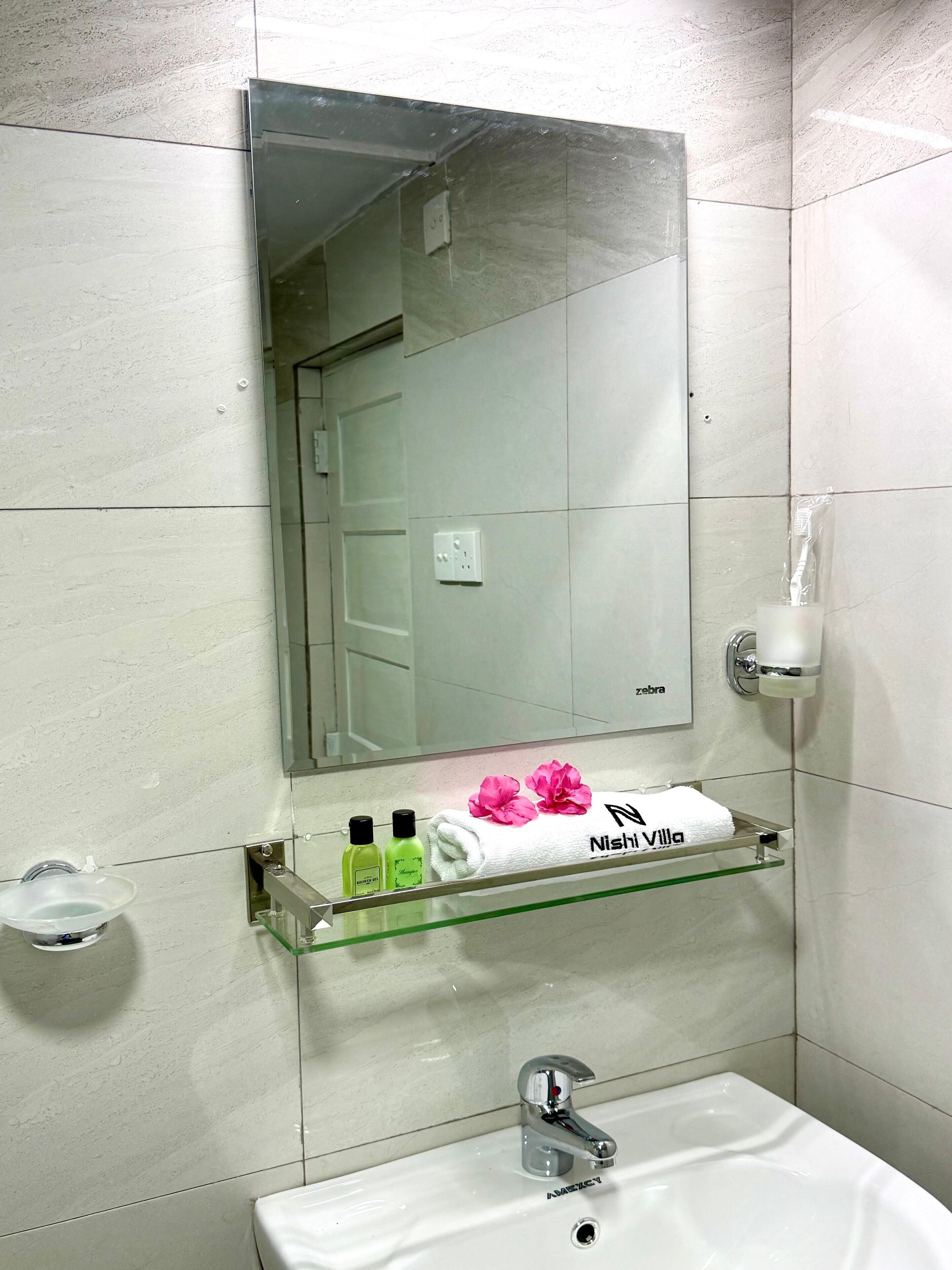 Un baño con espejo, lavamanos, repisa de vidrio con artículos de aseo y toallero con toalla y flores.