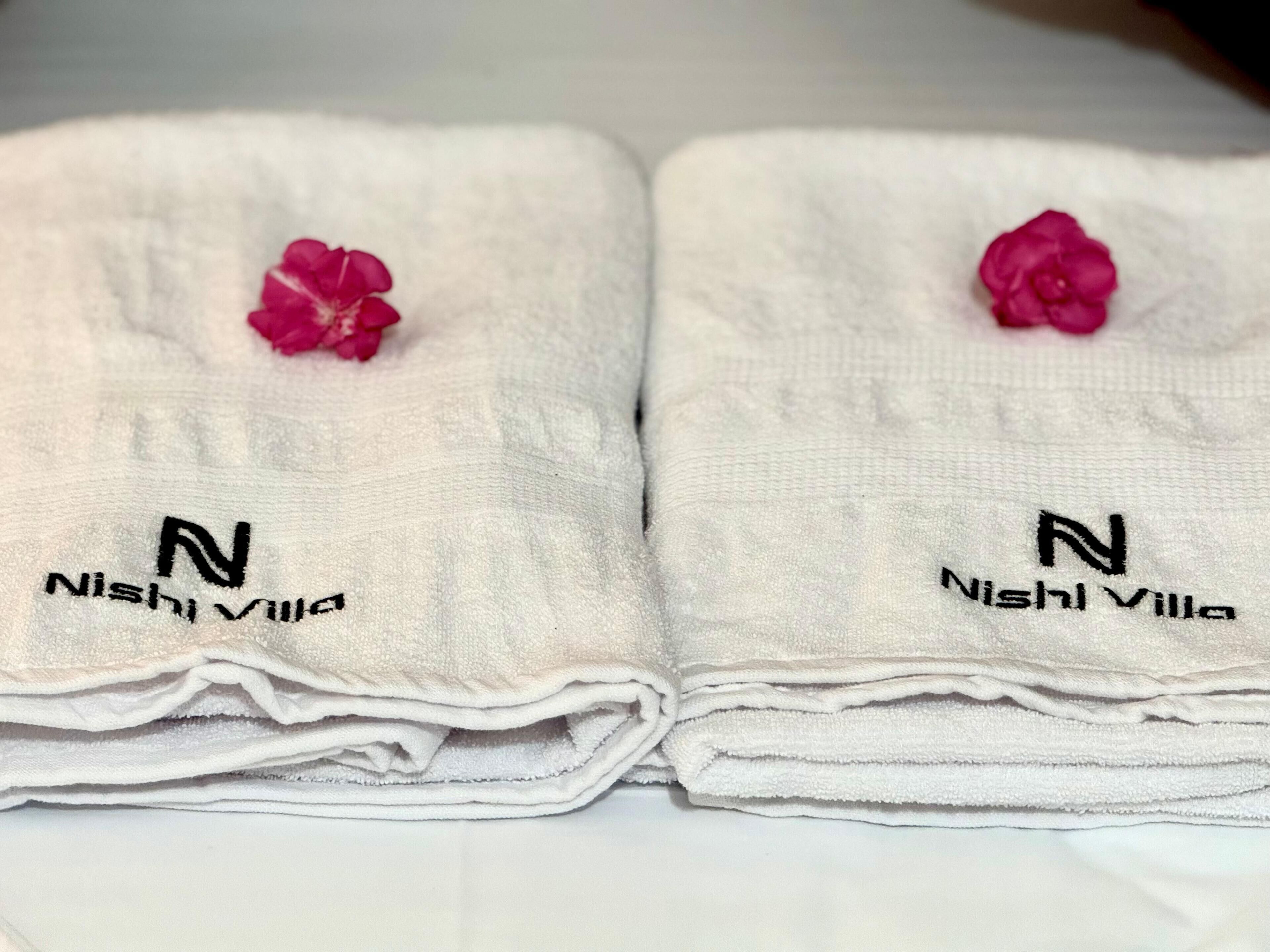 Dos toallas blancas dobladas con pulcritud, con el logo "Nishi Villa" y una flor rosa encima.