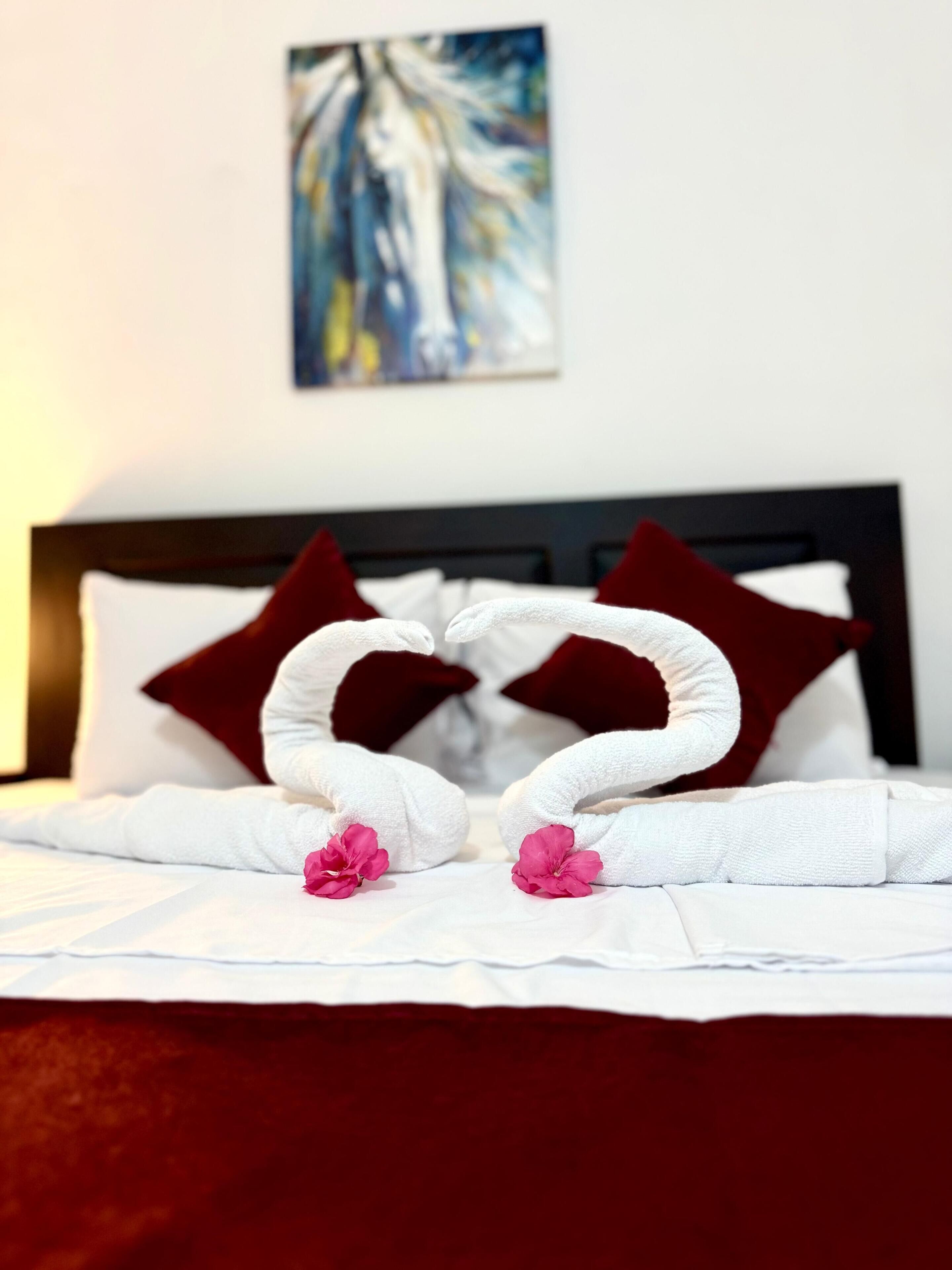 Una cama bien hecha con sábanas blancas, dos toallas con forma de corazón y dos flores rosadas.