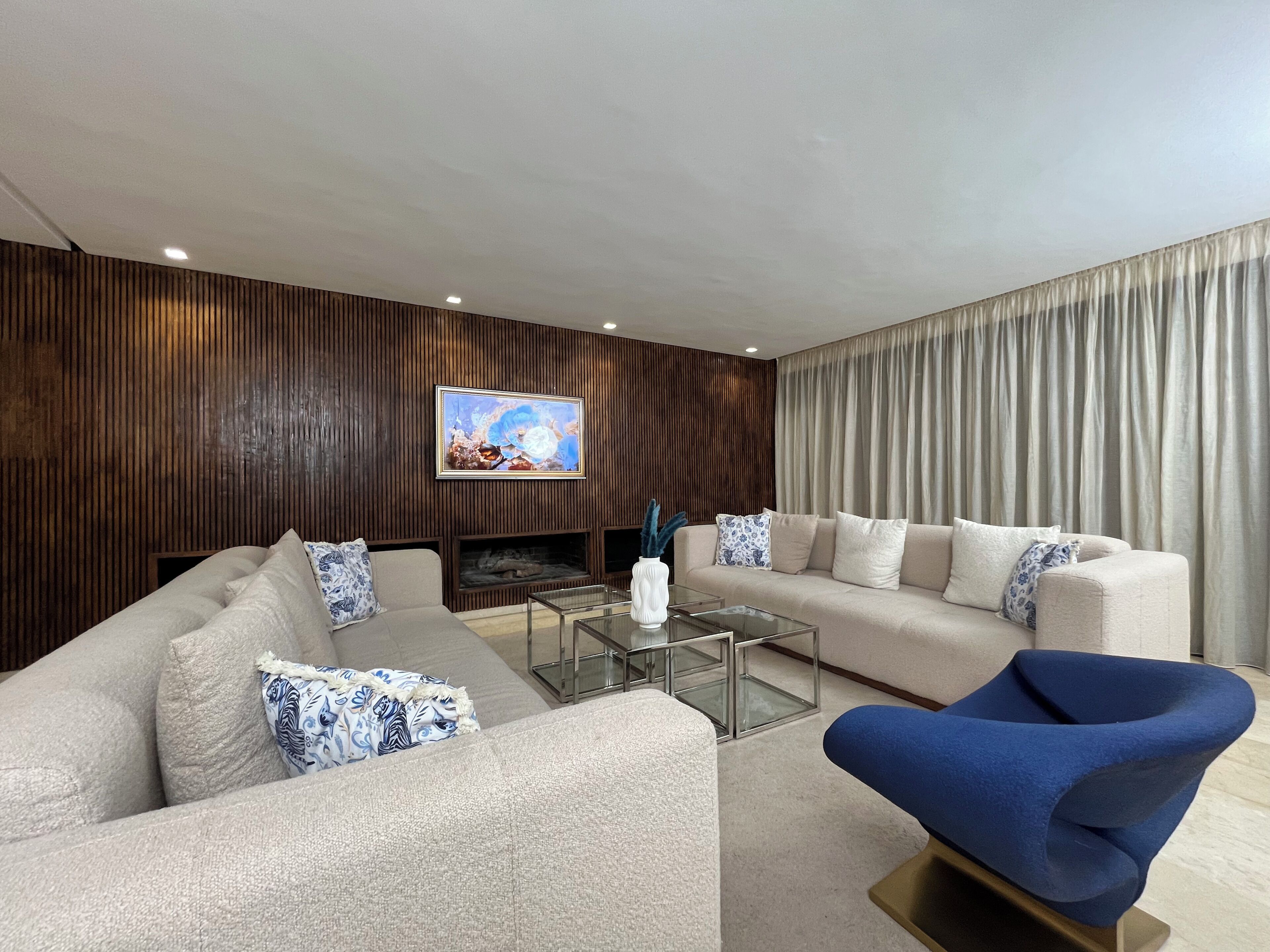 Una sala moderna con sofá, sillón y mesa de centro, un televisor de pantalla plana montado en la pared y un gran ventanal con cortinas.