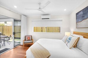 Un dormitorio con cama, ventilador de techo, equipo de aire acondicionado, una silla de madera, una mesa y vistas al exterior.