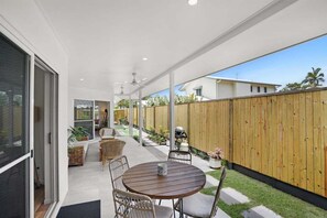 Un patio cubierto con una mesa redonda y sillas, una valla de madera y vista a una casa al fondo.